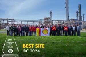 Group of Shell Sarnia Manufacturing Center team members standing together on a grassy field in front of an industrial plant, holding a Shell flag, with the IPA Best Site Award trophy displayed prominently in the foreground. The award recognizes their achievement in being named the Best Site for 2024.