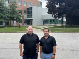 Paul Croft and Mike Thompson, Vice Presidents at Wolverine Contracting and Engineering, are pictured outside the Western Sarnia-Lambton Research Park. They are standing side by side in front of the building, wearing black polo shirts and smiling at the camera. The building features modern architecture with a mix of brick and metal siding, surrounded by greenery and trees.