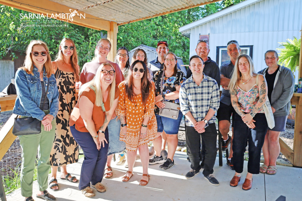 A group of twelve people standing together under a covered outdoor area, smiling towards the camera. The group consists of a diverse mix of men and women, all casually dressed. 